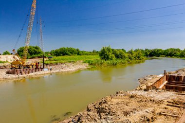 Suyun karşısında sarı renkli taşınabilir vinç ve pnömatik çekiç var. İnşaat alanındaki köprü temeli için nehir kıyısı boyunca inşa edilmiş metal yığınları. 