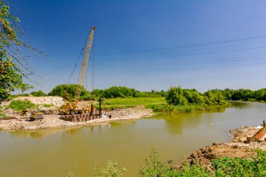 Suyun ötesine bakan sarı renkli taşınabilir vinç ve pnömatik çekiç. İnşaat alanındaki köprü temeli için nehir kıyısı boyunca inşa edilmiş metal yığınları. 