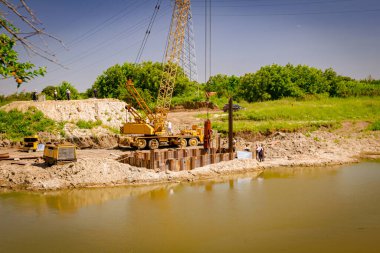 Suyun ötesine bakan sarı renkli taşınabilir vinç ve pnömatik çekiç. İnşaat alanındaki köprü temeli için nehir kıyısı boyunca inşa edilmiş metal yığınları. 