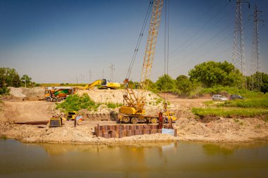 Suyun karşısında sarı renkli taşınabilir vinç ve pnömatik çekiç var. İnşaat alanındaki köprü temeli için nehir kıyısı boyunca inşa edilmiş metal yığınları. 