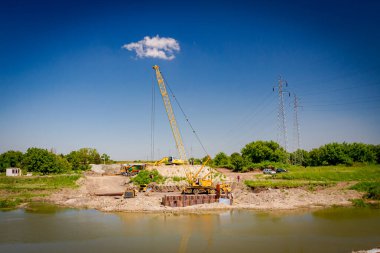 Suyun karşısında sarı renkli taşınabilir vinç ve pnömatik çekiç var. İnşaat alanındaki köprü temeli için nehir kıyısı boyunca inşa edilmiş metal yığınları. 