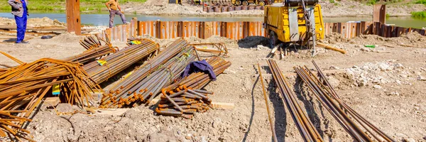 Nehir kıyısı boyunca demir yığınları döşeyip köprü temeli oluşturduk. Arka planda seçilmiş destek çubukları ve sarı mobil pnömatik kompresör paketleri var. İnşaat alanı