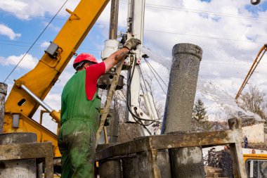 Rigger, kasklı işçi hortumu suyla yıkamak için kullanıyor, kanalı dikey şarj etmek için pompalıyor, nehir kıyısındaki köprü temeline taze beton döküyor..