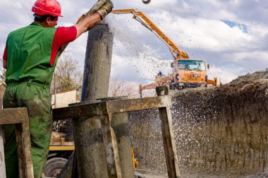 Rigger, kasklı işçi hortumu suyla yıkamak için kullanıyor, kanalı dikey şarj etmek için pompalıyor, nehir kıyısındaki köprü temeline taze beton döküyor..