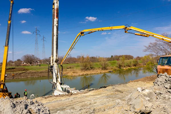 Köprü temellerine taze beton dökülüyor, nehir kıyısında yapım aşamasında. Mobil vinç ve kamyon pompası çalışıyor. Kule sondaj makinesi ayakta, zemini delmek için büyük ekipman var..