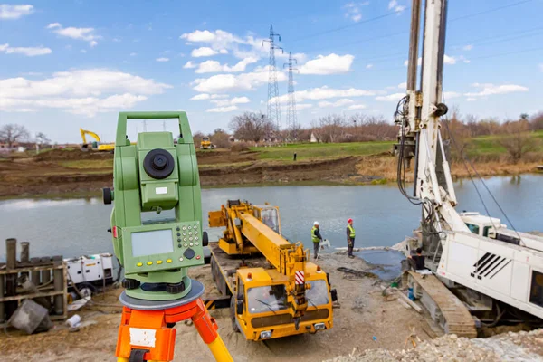 Surveyor instrument is for measuring level on building site. Mobile crane and drilling machine for drill into the ground are operating on bridge foundation at river coast.