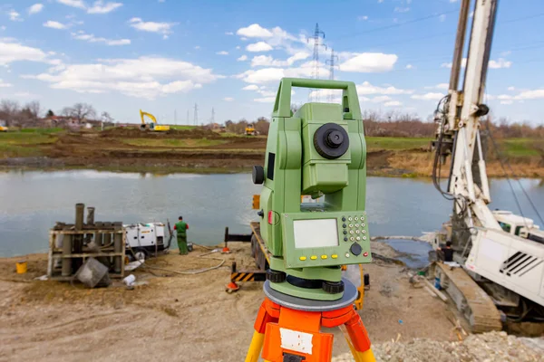 Surveyor instrument is for measuring level on building site. Mobile crane and drilling machine for drill into the ground are operating on bridge foundation at river coast.