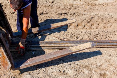 Koruyucu eldivenli işçiler halatı forklifte bağlar, kargoyu bağlar, uzun paslı armatür çubukları taşır, inşaat alanında yük taşır.. 