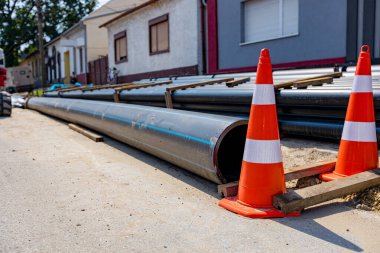 Birkaç turuncu trafik konisi, sokaklara istiflenmiş ve inşaat alanındaki siperlere yerleştirilmeye hazır yeni plastik su boruları paketinin yanında dikkat sembolüdür..