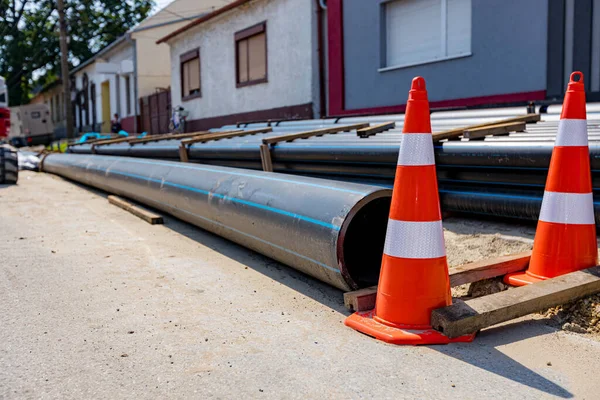 Several orange traffic cones are symbols of caution next to package of new plastic water pipes stacked on the street, ready to be placed in trench at building site.