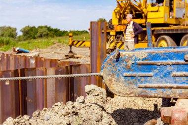 İki kancalı ve metal kollu el teli vinç, arka planda hareketli vinç ve metal kazıklarla çevrili siper, köprü yapım aşamasında..