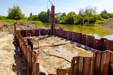 Pnömatik çekiçli mobil vinç kamyonu inşaat alanındaki nehir kıyısı boyunca köprü temeline metal kazıklar kuruyor. İşçi halat çekme kolu ile çelik sütunları doğru yöne koymayı başarıyor..