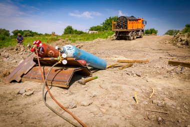 Kırmızı ve mavi kaynak ekipmanları, vanaları ve basınç göstergeleriyle oksijen ve nitrojen tankları, inşaat alanındaki metal kazıklara asetilen gaz silindirleri, rezervuarlı kamyon arka planda.