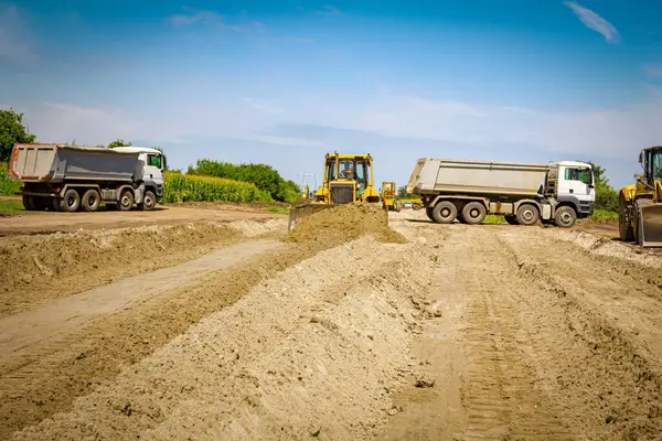 Tırtıllı toprak taşıyıcı, park halindeki kamyonlarda toprağı hareket ettiriyor ve arka planda başka bir buldozer var..