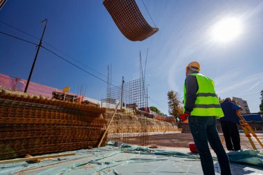 Güvenlik miğferi olan işçiler, inşaat alanının üzerinde beton takviyesi için paslı dikdörtgen çelik yığını taşırken vinçle teslimatı bekliyorlar..