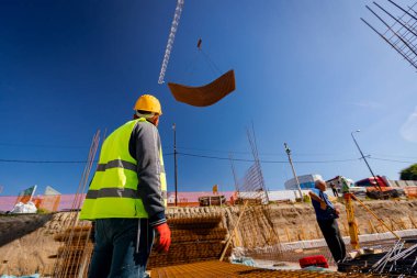 Güvenlik miğferi olan işçiler, inşaat alanının üzerinde beton takviyesi için paslı dikdörtgen çelik yığını taşırken vinçle teslimatı bekliyorlar..