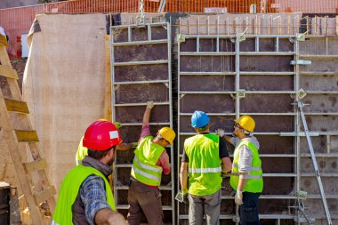 Güvenlik yeleği ve sarı miğferli inşaat işçilerinden oluşan bir ekip olarak, yüksek ahşap sökülebilir kalıpları elle monte ederlerken, amirin arkasından bakın..
