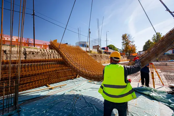 Güvenlik miğferli işçiler vinçin yerden kalkmasına yardım ediyor, dengeyi ve yönü koruyorlar, paslı dikdörtgen çelik desteğini inşaat alanının üzerine taşırken.