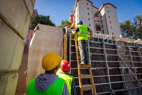 Güvenlik yeleği ve sarı miğferleri olan inşaat işçilerinden oluşan bir ekibin arkasından yüksek ahşap sökülebilir kalıpları elle monte edişini izleyin. İçlerinden biri merdivenlere tırmandı..