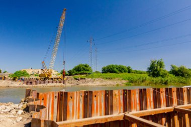 İnşaat alanındaki köprü temeli için nehir kıyısı boyunca inşa edilmiş paslı metal yığınları. Suyun karşısında sarı renkli, pnömatik çekiçli taşınabilir vinç var..