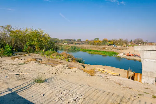 stock image Large modern gray concrete pillar in foundation surrounded by metal piles. Planks, beams placed on ground, wood profiles for building bridge molds, reinforced pole under construction is across river.