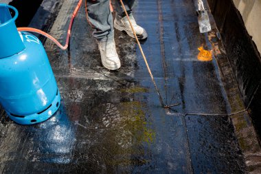 İşçi, propan pürmüz kullanılarak beton üzerine ziftle su yalıtımı kurulumunu ısıtıyor, eritiyor ve çalışma araçlarıyla yayılıyor. İnşaat halindeki konut binası su yalıtımı