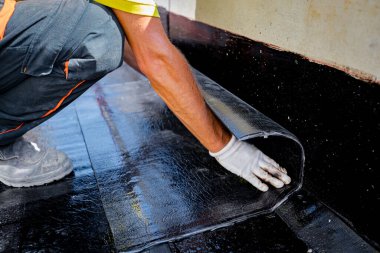 Eldivenli işçiler erimeye, elle yerleştirilmeye ve açılmaya hazırlanıyorlar. Su geçirmez döşeme betonun üzerinde. İnşaat halindeki konut binası su yalıtımı