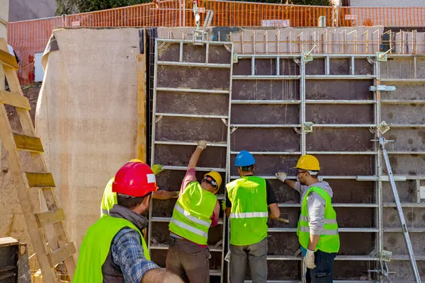 stock image View from behind on supervisor as overseeing team of construction workers with safety vest and yellow helmets as they manually assembly tall wooden demountable big mold.