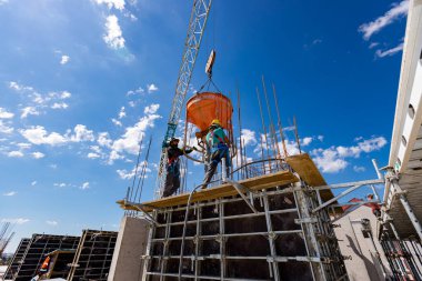İnşaat işçileri konik metal kovaları boşaltıyorlar, taşınıyor ve beton karışımını yüksek tutuyorlar, binanın sütunları için dikey kalıba beton döküyorlar, koni tankını taşırken vinç kullanıyorlar..