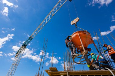 İnşaat işçileri konik metal kovaları boşaltıyorlar, taşınıyor ve beton karışımını yüksek tutuyorlar, binanın sütunları için dikey kalıba beton döküyorlar, koni tankını taşırken vinç kullanıyorlar..