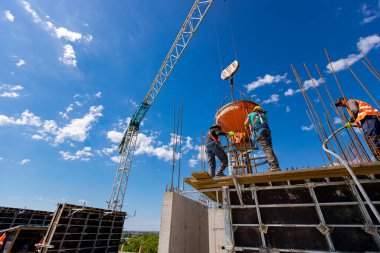 İnşaat işçileri konik metal kovaları boşaltıyorlar, binanın sütunları için yüksek dikey kalıplara beton döküyorlar, koni tankı taşırken vinç kullanıyorlar. İşçi güç aracı kompakt beton kullanıyor.