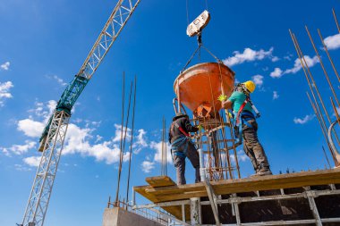 İnşaat işçileri konik metal kovaları boşaltıyorlar, taşınıyor ve beton karışımını yüksek tutuyorlar, binanın sütunları için dikey kalıba beton döküyorlar, koni tankını taşırken vinç kullanıyorlar..