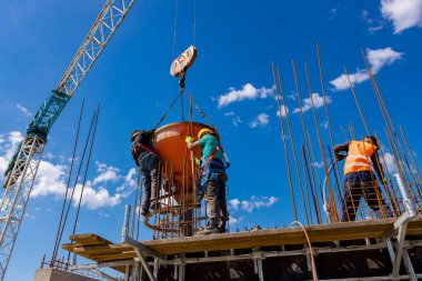 İnşaat işçileri konik metal kovaları boşaltıyorlar, binanın sütunları için yüksek dikey kalıplara beton döküyorlar, koni tankı taşırken vinç kullanıyorlar. İşçi güç aracı kompakt beton kullanıyor.
