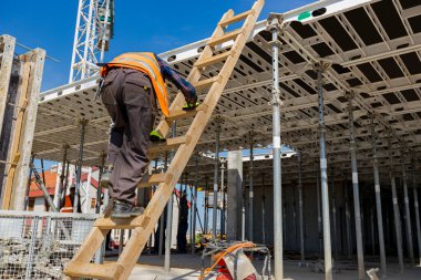 Güvenlik yeleği ve miğferi olan inşaat işçisinin arkasından ahşap merdivenlere tırmanırken inşaat halindeki bir sonraki kata bakın..