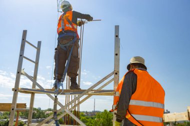 Denetmen, ustabaşı beton sütun için takviye iskeleti yapmak için armatür bağlayan işçiye göz kulak olur. Düşme tutuklaması cihazı taşır. Emniyet kemeri de iskeleye zımparalanır.. 