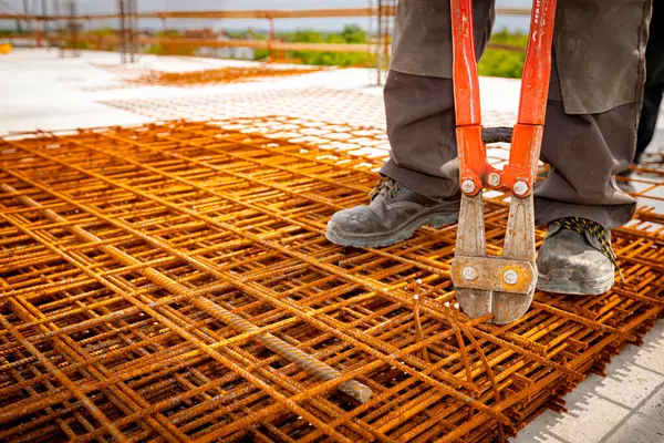 stock image Worker is standing on pile of rusty rectangle steel, reinforcement for concrete as cutting reinforcement grid, mesh with bolt cutters at building site.