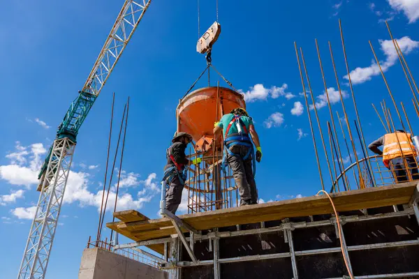 İnşaat işçileri konik metal kovaları boşaltıyorlar, binanın sütunları için yüksek dikey kalıplara beton döküyorlar, koni tankı taşırken vinç kullanıyorlar. İşçi güç aracı kompakt beton kullanıyor.