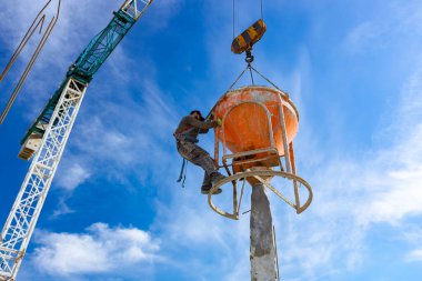 İnşaat işçileri konik metal kovaları boşaltıyorlar, taşınıyor ve beton karışımını yüksek tutuyorlar, binanın sütunları için dikey kalıba beton döküyorlar, koni tankını taşırken vinç kullanıyorlar..