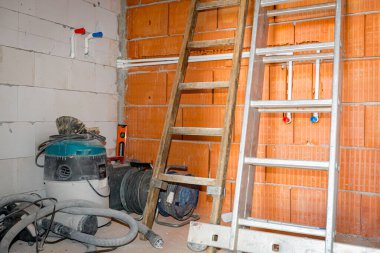 Ahşap ve metal merdivenler, kırmızı bloklardan yapılmış duvara yaslanmış, yapım aşamasındaki odanın köşesine yerleştirilmiş, elektrikli süpürge ve makaralı birkaç elektrik uzatma kablosu var..