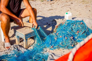 Bir balıkçı sahilde bir sandalyede oturur ve deniz kabuklarından balık ağını temizler, onları tahta bir tahtayla ezer..