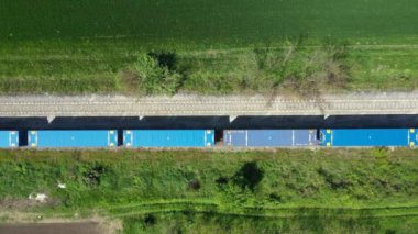 Uzun kompozisyonun üstünde, kargo konteynırlarından oluşan demiryolu yük treni tarımsal alanların çeşitli ekili alanlarından geçiyor..