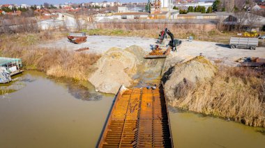 Yukarıda, kazıcı mücadele ediyor, eski yük gemisinin bir kısmını, mavnayı, nehir kıyısından, kasıtlı olarak, parça parça sökmeye, paslı metali, geri dönüşüm için kesmeye çalışıyor..