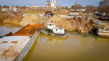 Above view over excavator, tow truck and tugboat as they pull out part of old freight vessel's bed, barge, at river coast, cassation, dismantling ship, metal for scarp, recycling. clipart