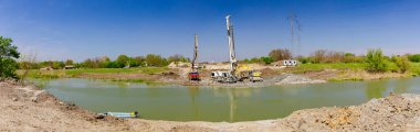 Panoramik manzara, endüstriyel kule sondaj makinesi, zemini delmek için büyük aletler. Pompalı kamyonet nehir kıyısındaki köprü temeli için taze beton döküyor..