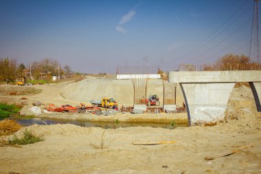Temelde metal yığınlarıyla çevrili büyük gri beton köprü sütunları. İnşaat halindeki güçlendirilmiş direk nehrin karşısında. Ekip çalışıyor.