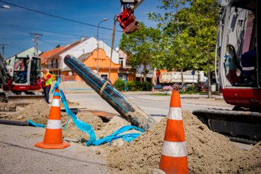 Beyaz floresan şeritli birkaç turuncu trafik konisi, su tedarik etmek için su tesisatı, plastik HDPE boru hattını tutmak için kayışlar kullanıldığı için kazıcının yanındaki uyarıyı işaret ediyor.. 