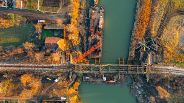 Above top view on workers are cutting old railroad bridge from cherry picker, with acetylene torch for recycling. Large crane on vessel, barge, hold scrap metal, docked at river coast under bridge. clipart