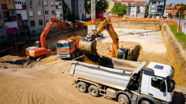 Kazı yaparken iki büyük kazıcının üstünde, yeni konut binası için kazı yapıyorlar ve birkaç çöp kamyonunu toprakla dolduruyorlar, inşaat alanında, proje devam ediyor.