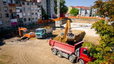 Kazı yaparken iki büyük kazıcının üstünde, yeni konut binası için kazı yapıyorlar ve birkaç çöp kamyonunu toprakla dolduruyorlar, inşaat alanında, proje devam ediyor.