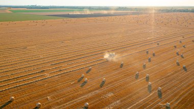 Manzaranın üstünde traktörün arka ışığı mercimeği fırına verirken, kamışı yuvarlayan ve tarımsal alanın üzerine yuvarlanmış bir balya tüküren makine, kapağı açar ve paketi dışarı atar..